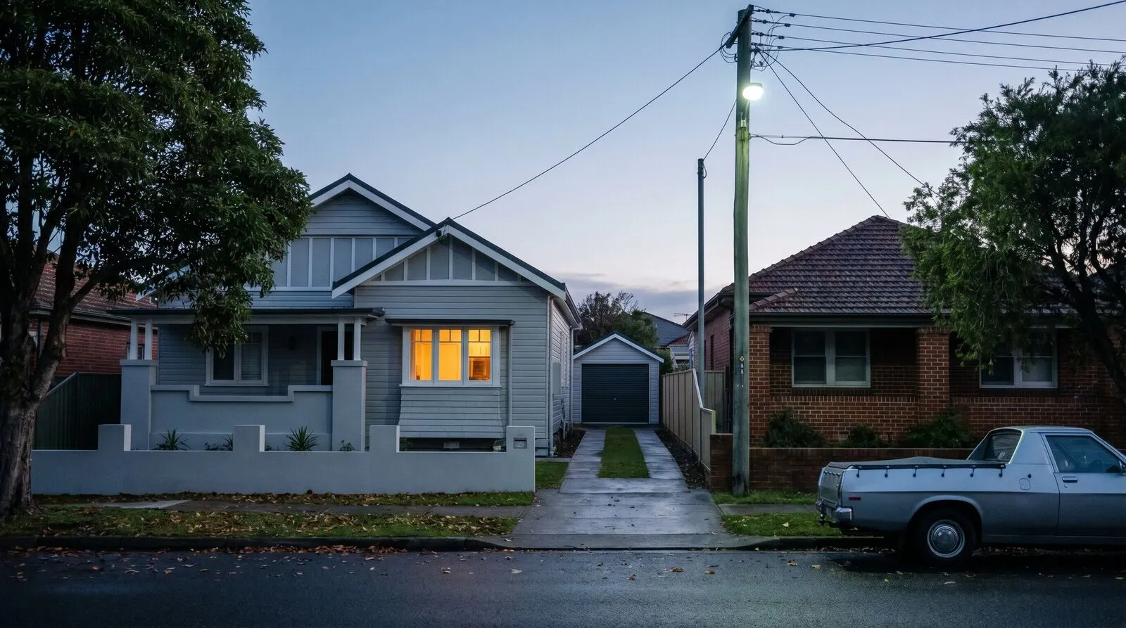 A New Lambton street before dawn: a bungalow with a side driveway, a ute at the kerb and one kitchen window lit